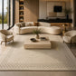 A large cream-colored area rug with a subtle geometric border pattern, styled under a bed in a modern, warm-toned bedroom with wood paneling and natural light.