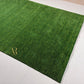 A rectangular green artificial turf or grass mat with a textured grass-like surface, laid out on a light-colored tiled floor. A gold monogram logo reading 'AR' is printed near one corner.