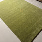 An olive green textured area rug with a small gold monogram logo in the lower right corner, displayed flat against a white background.