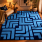 A large area rug with a bold geometric maze-like pattern in light blue and dark navy blue, styled in a warm, modern living room setting.