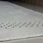 A close-up of a light beige or off-white rug with a textured, looped pile and rows of small dark tufted accents, shown on a glossy floor.