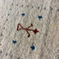 Close-up of a textured area rug with a cream-colored base, featuring a central rust-brown abstract symbol and scattered blue and gray motifs.