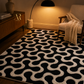 A black and cream wavy pattern area rug styled in a warm, modern living room setting next to a beige armchair and a lit floor lamp.