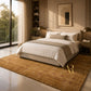 A modern bedroom scene featuring a large, plush shag area rug in a warm golden-brown color, styled under a neatly made bed with white and beige linens. The room is illuminated by warm natural light from a large window on the left, showcasing a sleek contemporary design with a bed frame, bedside tables, and lamps.