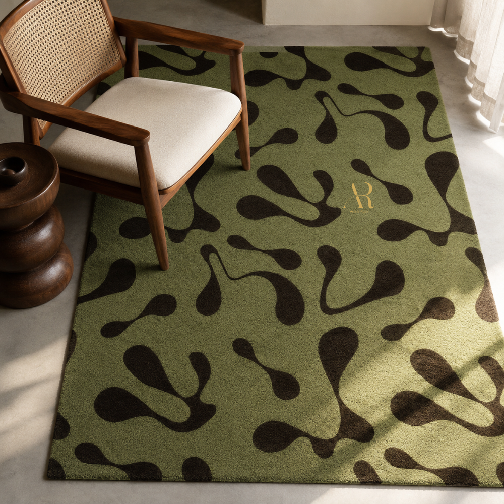 An olive green area rug with a dark brown abstract organic pattern and a gold monogram, styled in a modern living room next to a wooden armchair.