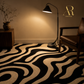 A black and beige abstract wavy patterned area rug in a styled living room setting with a floor lamp, wooden side table, and leather armchair. The brand logo 'Artisa Rugs' is visible in the top right corner.