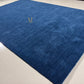 A royal blue area rug with a small gold embroidered monogram logo, shown laid out on a light-colored tiled floor.