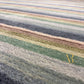 A close-up of a textured area rug with horizontal stripes in muted tones of gray, sage green, beige, and brown, featuring a gold 'R' logo in the lower right corner.