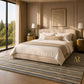A styled bedroom interior featuring a cream upholstered bed with white and beige bedding, flanked by gold bedside tables and lamps, set on a striped blue, beige, and gray rug. A large window on the left shows a forest view.