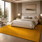 A large mustard yellow area rug with a geometric border pattern is styled in a modern, neutral-toned bedroom with a bed, nightstands, and a window view.