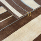 A close-up of a striped area rug with diagonal bands of dark brown, cream, and light blue, featuring a gold 'AR MONASTOR' logo watermark.