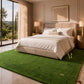 A luxury bedroom featuring a large green turf-style area rug with gold monogram details, styled with a beige upholstered bed, white bedding, and warm ambient lighting.