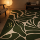 A green and beige leaf-patterned area rug styled in a warm, modern living room setting with a wooden floor, a lit floor lamp, and a wooden console table in the background. The brand logo 'Artina Rugs' is visible in gold near the bottom left of the rug.