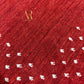 A close-up of a red textured fabric with a diagonal ribbed pattern and scattered small white motifs. A gold monogram logo reading 'AR' with smaller text beneath appears near the upper left.