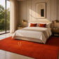 A styled bedroom scene featuring a large rust-red area rug with a gold monogram logo, under a bed dressed in white and beige linens with two orange accent pillows. The room has warm beige walls, modern bedside tables with gold lamps, and a large window looking out onto green trees.