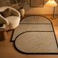 A modern geometric area rug with a slatted natural fiber pattern and black trim, styled in a warm, contemporary living room next to a cream-colored armchair and a lit floor lamp.