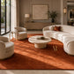 A large rust-orange area rug in a modern living room setting, styled with a curved cream sofa, two matching armchairs, a round stone coffee table, and tasteful decor.