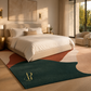 A modern bedroom scene with a large abstract area rug in dark teal, terracotta, cream, and blush tones. The rug is styled on a light wood floor around a bed with white linens and beige bedding.