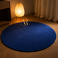 A round deep blue area rug on a wooden floor in a warmly lit room, with a glowing floor lamp and curtains in the background. A stylized gold 'R' logo with 'Artist Rug' text is printed near the center of the rug.