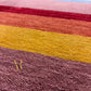 A rectangular area rug with vertical stripes in multiple colors including brown, blue, red, orange, pink, yellow, green, and light blue, featuring a gold embroidered monogram logo in the bottom left corner.