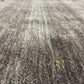 A close-up of a textured area rug with a shaggy, looped pile in a mottled gray and beige colorway, featuring a subtle gold monogram logo in the lower right corner.
