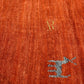 A close-up of a textured orange woven fabric with a small gray embroidered motif in the lower right and a faint gold monogram near the upper center.