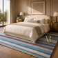 A styled bedroom scene showing a bed dressed in cream bedding and a beige throw, placed on a striped area rug with blue, red, and cream bands. The room has warm wood flooring, gold bedside tables and lamps, curtains, and a large window.
