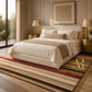 A styled bedroom scene showing a bed dressed in white bedding with beige accents, placed on a striped area rug in a warm neutral room. Large windows, gold bedside tables and lamps, and framed wall art are visible.