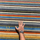 A rectangular area rug with a vibrant multicolor horizontal stripe pattern, featuring a small gold embroidered monogram logo in the upper left corner, set against a plain white background.