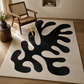A large rectangular area rug with an abstract black organic pattern on a warm off-white background, styled in a modern living room setting with a wooden armchair, side table, and plant.