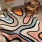 A modern abstract area rug with flowing wavy lines in beige, blush pink, light blue, orange, and black, styled on a light wood floor next to a wooden armchair.