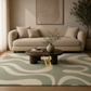 A styled living room scene featuring a beige sofa, a round dark wood coffee table, and a large area rug with a wavy abstract pattern in sage green and cream. A gold watermark logo for 'Artisan Rug' is visible on the rug.