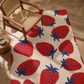 A rectangular area rug with a cream background and a bold pattern of large red strawberries with dark blue tops, styled in a room with a wooden armchair and a small round wooden side table.