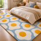 A plush area rug with a repeating fried-egg pattern in light blue, cream, and mustard yellow, styled on a light wood floor in a bright, modern bedroom next to a bed with neutral bedding.