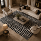A large rectangular area rug with a bold black and off-white geometric pattern, styled in a luxurious, high-end living room with a tufted sofa, armchairs, and a marble coffee table.