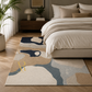 A modern abstract area rug with beige, navy, and slate blue organic shapes, styled on a light wood floor in a bedroom next to a bed with neutral bedding.