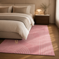 A pink area rug with a white wavy geometric pattern and a small gold monogram, styled on a wooden floor next to a bed in a modern, warm-toned bedroom.