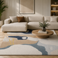 A modern beige sectional sofa in a styled living room, placed on a large abstract area rug with blue, beige, and cream tones. A round wooden coffee table with a vase of greenery sits in front of the sofa, and a plant and shelving are visible in the background.