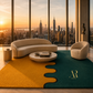A modern living room scene with a curved cream sofa, a matching armchair, a round coffee table, and a large area rug in mustard yellow and deep teal with a wavy seam design. The room is set in front of floor-to-ceiling windows overlooking a city skyline at sunset.