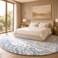 A large round area rug with a wavy abstract pattern in light blue, beige, and cream, styled in a modern bedroom setting with a bed, nightstands, and large windows.