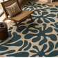A large area rug with an abstract organic leaf-like pattern in dark teal and warm beige, styled in a bright room with a wooden chair, a throw pillow, and a small side table.