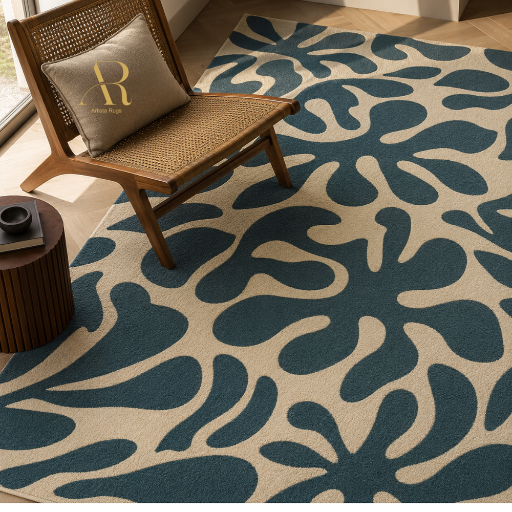 A large area rug with an abstract organic leaf-like pattern in dark teal and warm beige, styled in a bright room with a wooden chair, a throw pillow, and a small side table.
