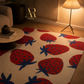 A cream-colored area rug with a bold pattern of large red strawberries and dark blue leafy tops, styled in a warm, modern living room setting with a floor lamp and wooden furniture.