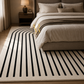 A modern area rug with a cream background and bold black curved and vertical stripe pattern, styled in a contemporary bedroom next to a bed.