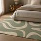 A modern area rug with a sage green and cream wavy abstract pattern, styled in a bedroom setting under a bed.
