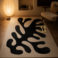 A rectangular area rug with an abstract black organic pattern on a warm cream background, styled in a cozy living room setting with a glowing floor lamp and a modern armchair.