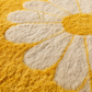 Close-up of a plush, textured rug featuring a large daisy pattern in mustard yellow and cream. A subtle gold monogram logo is visible in the center.