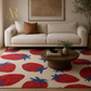 A styled living room scene featuring a cream-colored sofa with a brown accent pillow, a round wooden coffee table, and a large cream rug decorated with a bold red and blue strawberry pattern. A gold 'Artsie Rouge' brand logo is visible in the top left corner.