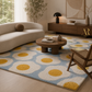 A modern living room scene featuring a plush area rug with a light blue background and large white and yellow egg-shaped patterns. The room is styled with a cream-colored curved sofa, a round wooden coffee table, a wooden chair, and a dark wood sideboard in the background.