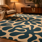 A large area rug with an abstract organic pattern in dark teal blue and warm cream, styled in a cozy living room setting next to a mid-century modern armchair and a wooden sideboard.